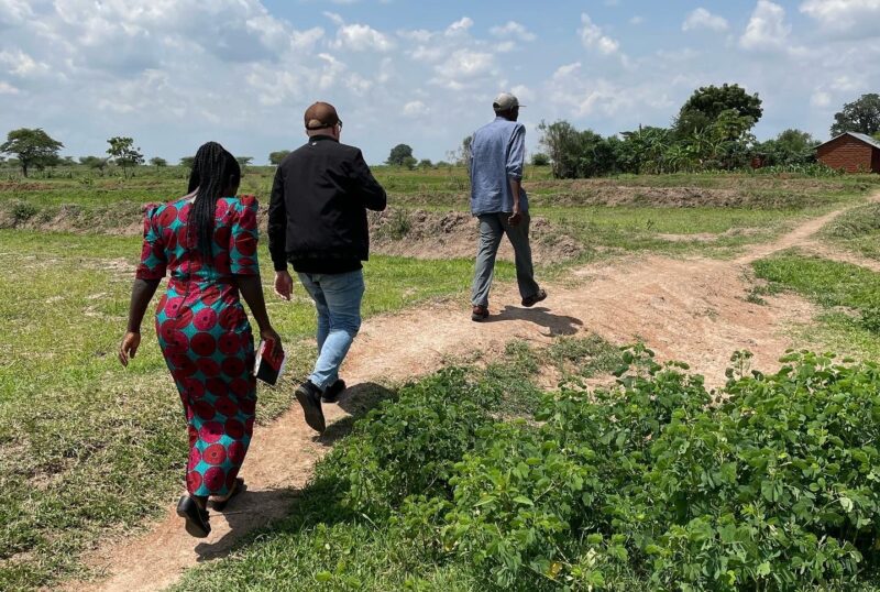 Three people walking on a dirt path through a green field under a cloudy sky.