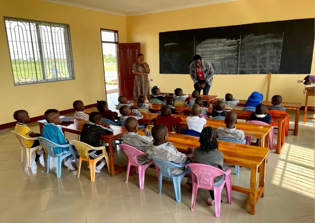 A classroom with young students seated at desks, two adults standing at the front near a blackboard.