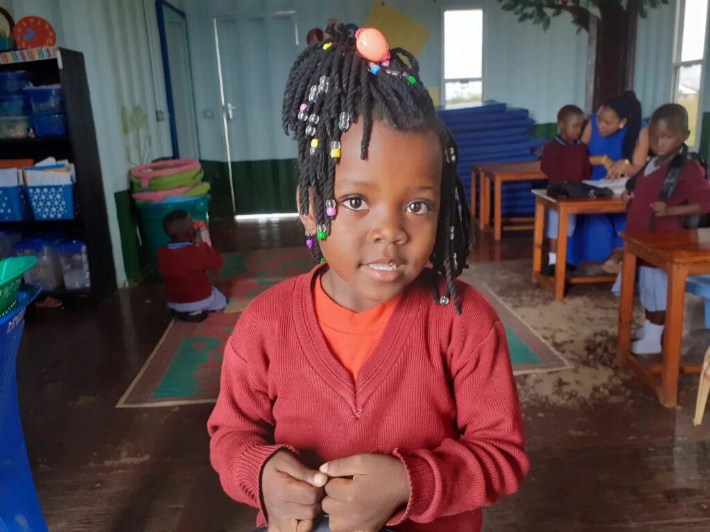 A young girl with beaded braids standing indoors, looking at the camera.