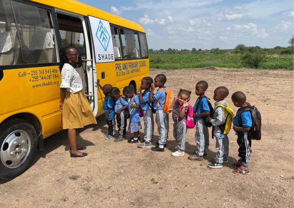 A group of young children standing in a line outside, waiting to board a yellow school bus labeled “SHADE.”