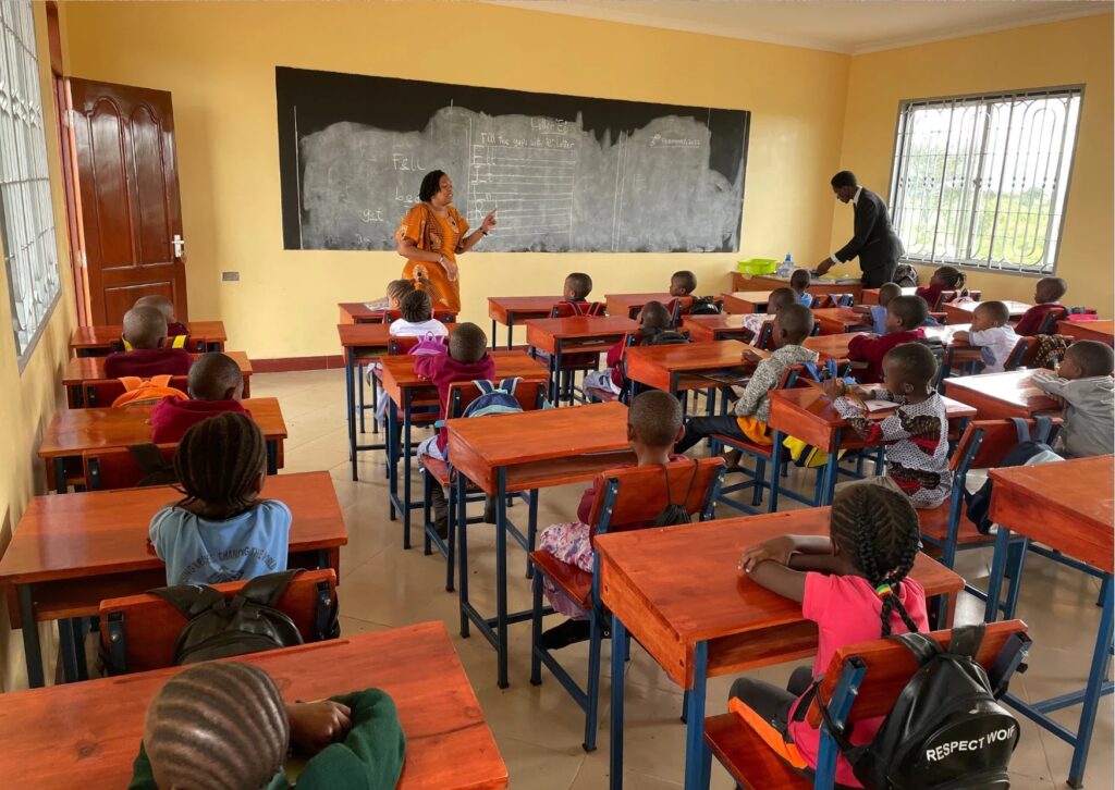 A classroom with young students seated at desks, a teacher at the front near a blackboard.