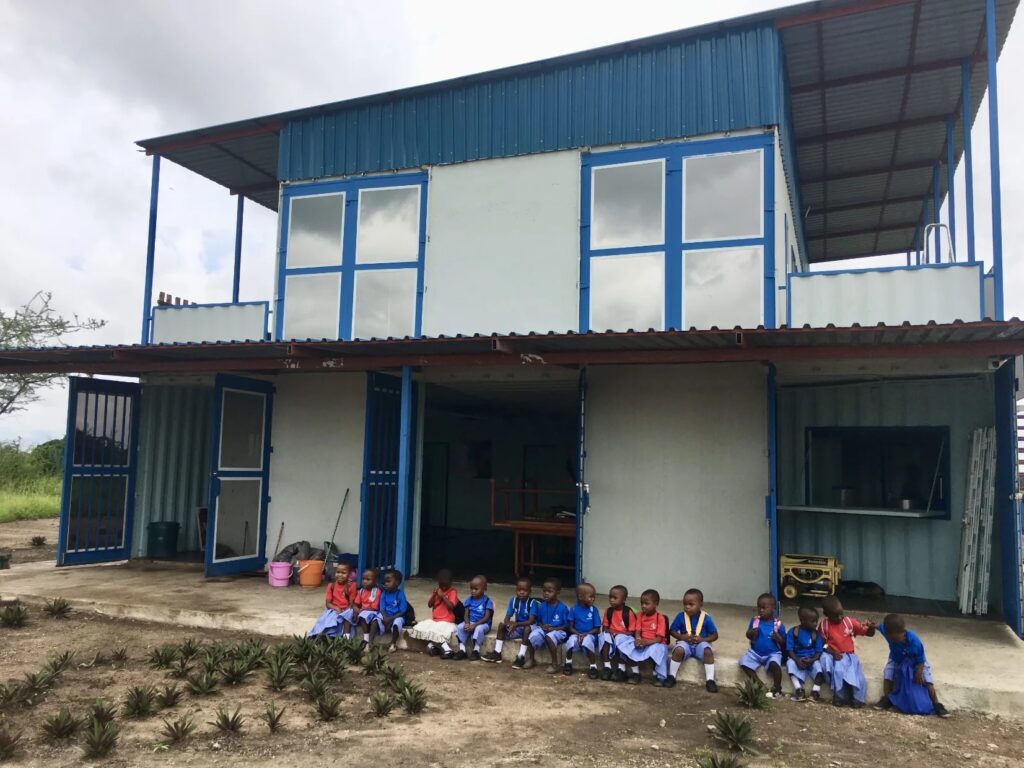 A group of young children in school uniforms sitting in front of a two-story building with blue accents.