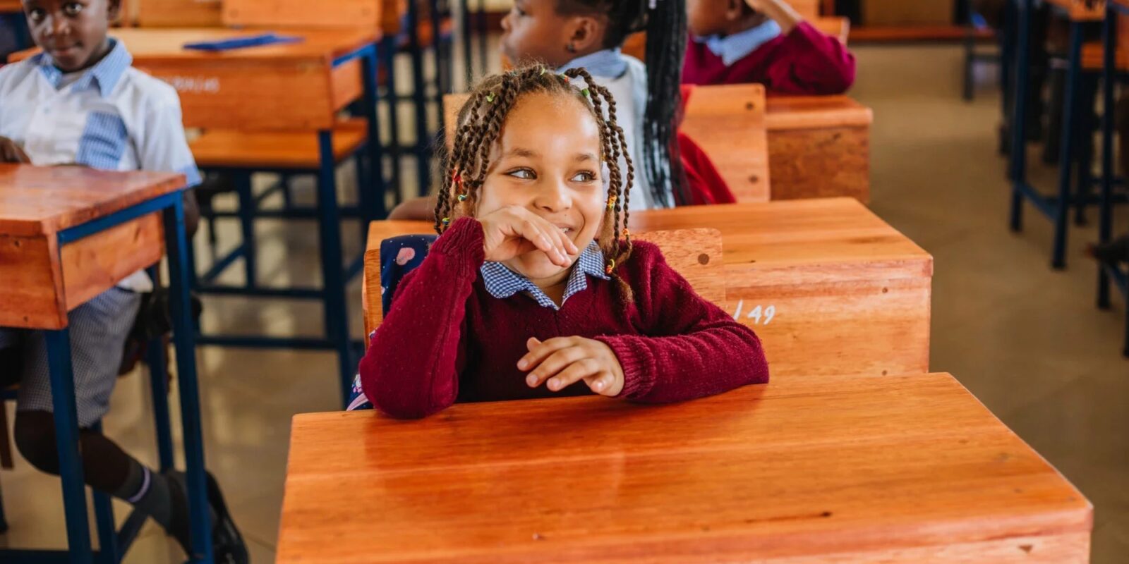 Children sitting at wooden desks in a classroom, with a girl in the foreground smiling.