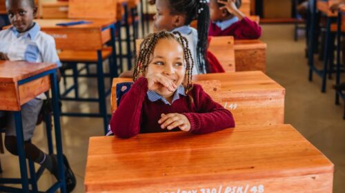 Children sitting at wooden desks in a classroom, with a girl in the foreground smiling.