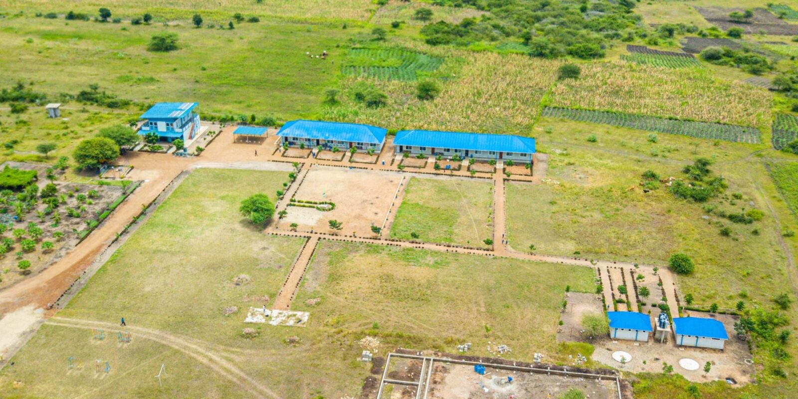 This aerial image depicts a large compound or campus set in a rural or semi-rural area. The compound has multiple buildings with blue roofs, likely used for various purposes such as classrooms, offices, or accommodations. The surrounding area is predominantly green, with patches of cultivated land, indicating agricultural activity. The layout appears well-organized, with pathways connecting the different buildings and sections of the compound. There are also some fenced areas and structures under construction, suggesting ongoing development or expansion. The overall setting reflects a tranquil environment with a focus on education and development.