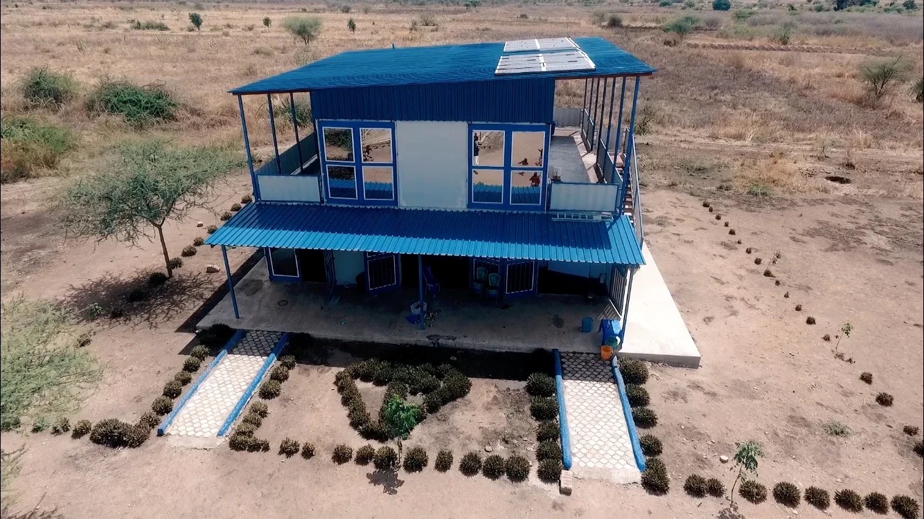 This image shows a two-story building with a blue roof, located in a rural or semi-rural area. The building appears to be well-constructed and maintained, with a large veranda on the ground floor and a balcony on the upper floor. The pathways leading to the building are lined with small plants, and the surrounding area is dry with sparse vegetation. The structure seems to be designed for multiple purposes, potentially serving as an administrative or residential facility within the community. The presence of solar panels on the roof suggests a focus on sustainability and renewable energy sources.