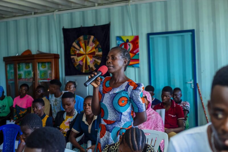 A woman in a colorful dress speaking into a microphone in front of a seated audience.