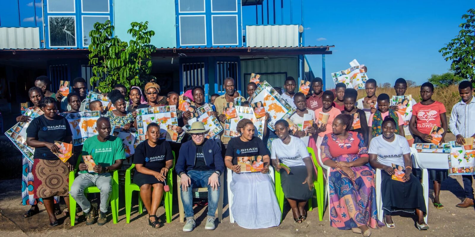 Group of people holding posters and booklets, standing and sitting in front of a blue building.