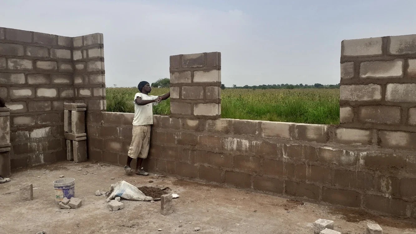 This image shows a construction worker building a wall with concrete blocks. The structure is in the early stages of construction, with partial walls already erected. The worker is positioned within the structure, actively placing or adjusting one of the blocks. The surrounding area appears to be a field or open land, suggesting that the construction is taking place in a rural or semi-rural environment. The worker is using basic construction tools and materials, and there is a bucket and some loose blocks and mortar on the ground nearby. The scene highlights the ongoing development and building efforts within the community.