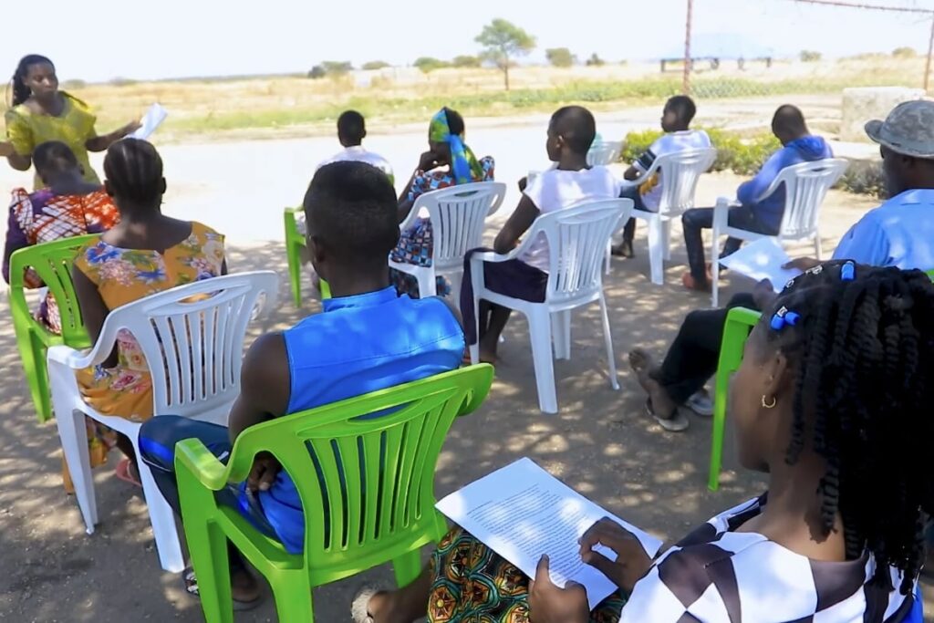 Group of people sitting on plastic chairs outdoors while a woman is speaking.