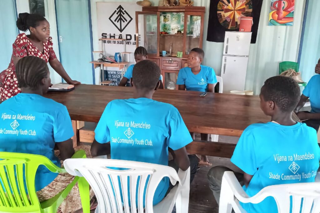 Group of young people in blue shirts sitting at a table inside a room, listening to a woman.