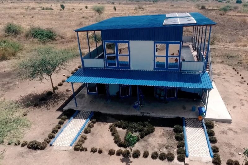 This image shows a two-story building with a blue roof, located in a rural or semi-rural area. The building appears to be well-constructed and maintained, with a large veranda on the ground floor and a balcony on the upper floor. The pathways leading to the building are lined with small plants, and the surrounding area is dry with sparse vegetation. The structure seems to be designed for multiple purposes, potentially serving as an administrative or residential facility within the community. The presence of solar panels on the roof suggests a focus on sustainability and renewable energy sources.