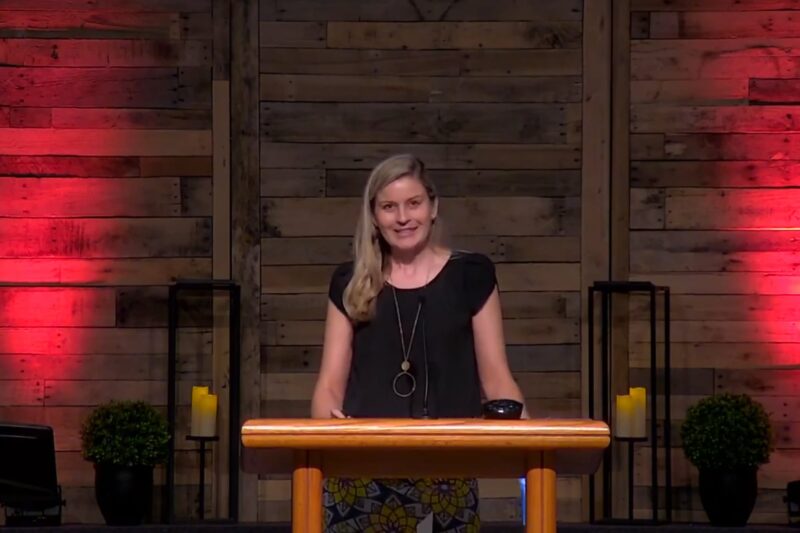 Woman speaking at a podium with a wooden background.