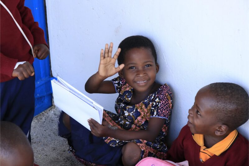 Smiling girl waving and holding papers while sitting next to another child.