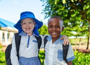 Two school children, one with albinism, smiling and standing with arms around each other.