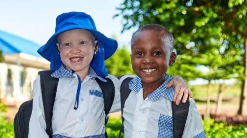 Two school children, one with albinism, smiling and standing with arms around each other.