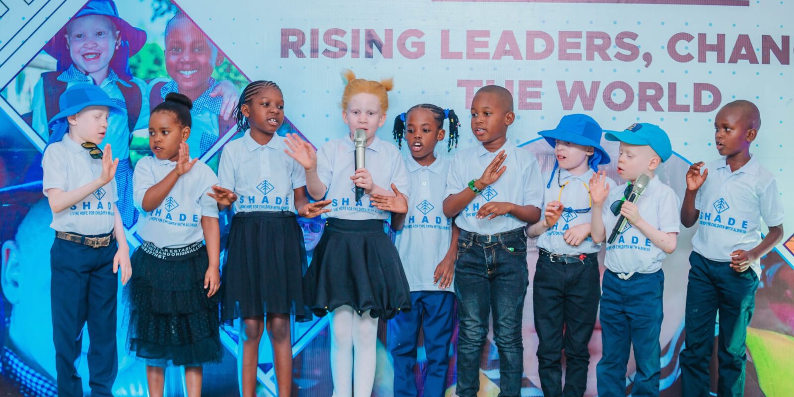 children standing on a stage wearing “SHADE” t-shirts, some holding microphones, participating in a performance or presentation.