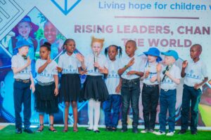 children standing on a stage wearing “SHADE” t-shirts, some holding microphones, participating in a performance or presentation.