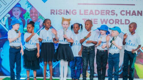 children standing on a stage wearing “SHADE” t-shirts, some holding microphones, participating in a performance or presentation.